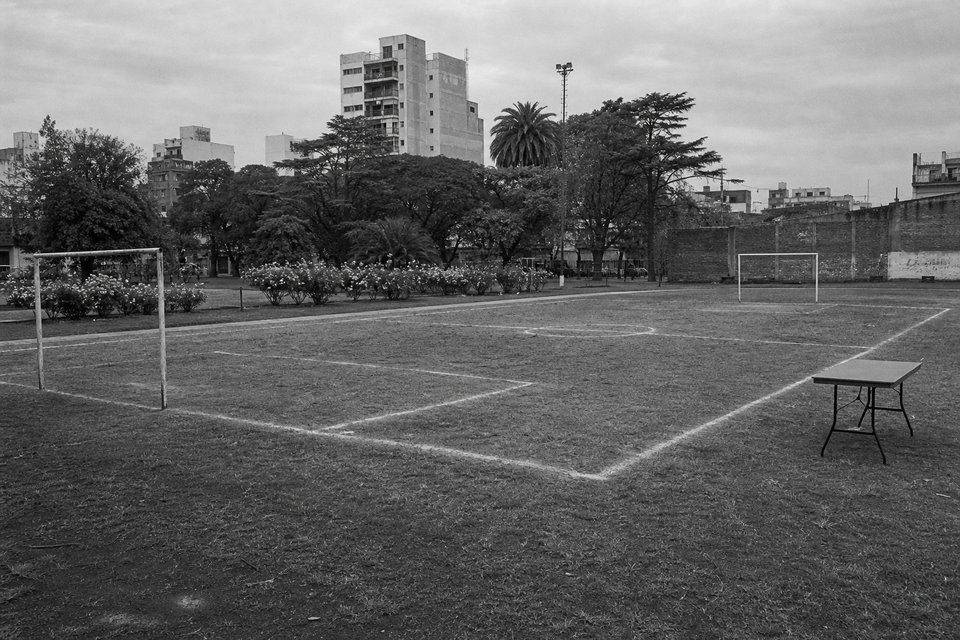 Cancha marcada con cal sobre el pasto de un parque urbano, con dos arcos sin red, rosales al fondo y una mesa plegable vacía a un costado