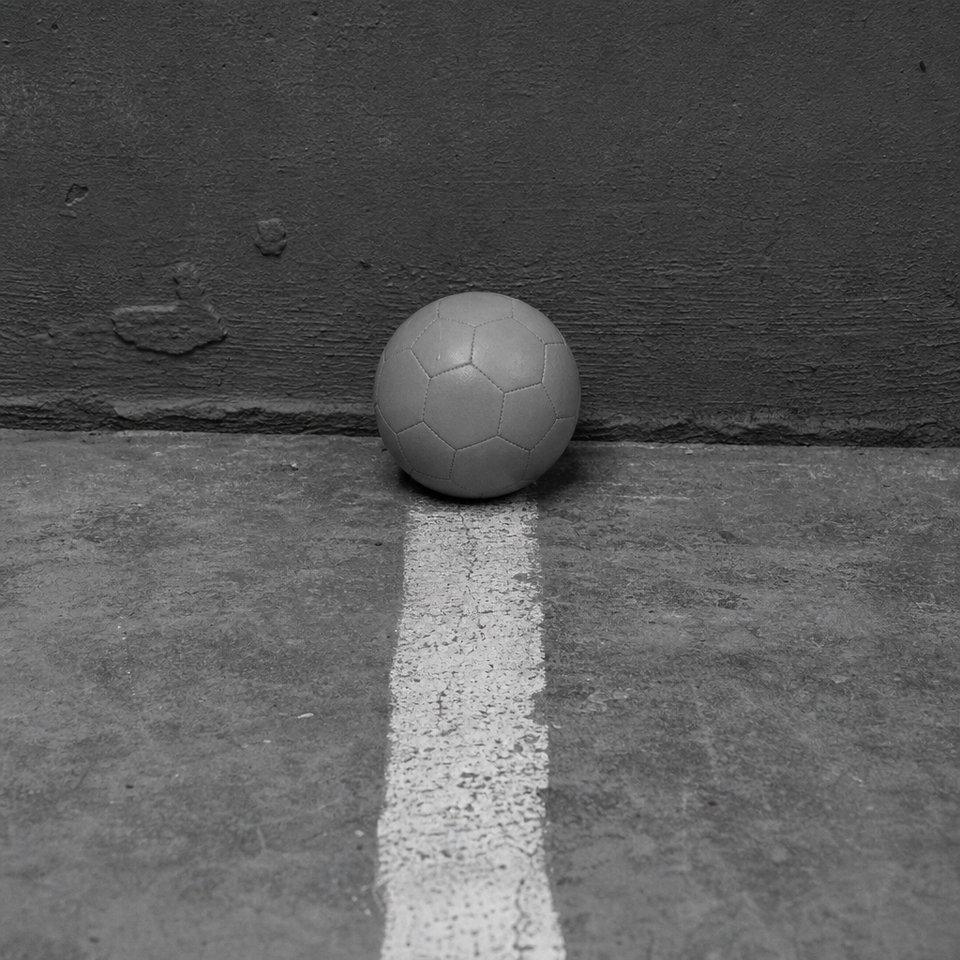 Pelota de futsal, más chica y de superficie mate, apoyada sobre una línea pintada en un piso duro y liso, con una pared al ras