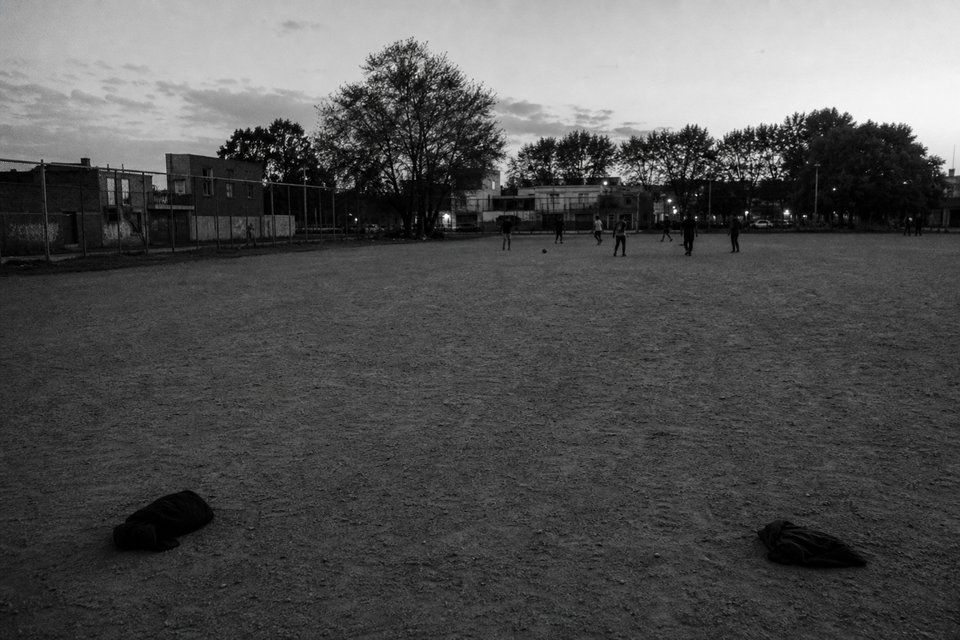 Cancha improvisada sobre tierra al atardecer, con camperas dobladas marcando un arco en primer plano y figuras lejanas al fondo