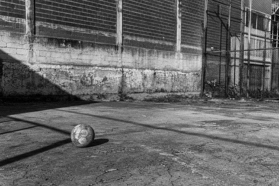 Pelota gastada sola sobre asfalto agrietado, con una sombra diagonal marcada cruzando el encuadre