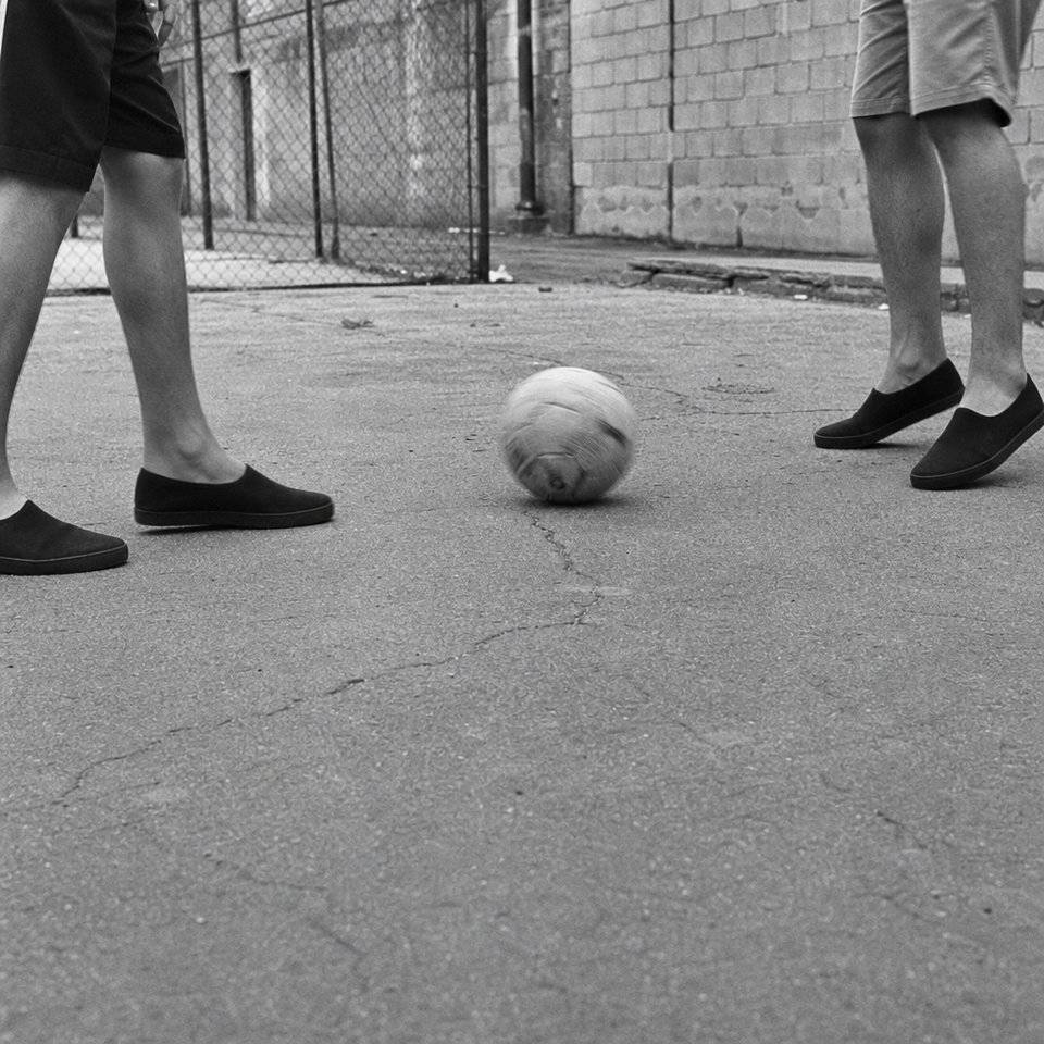 Dos jugadores enfrentados sobre asfalto, vistos de la rodilla hacia abajo, con la pelota rodando entre ellos en pleno pase